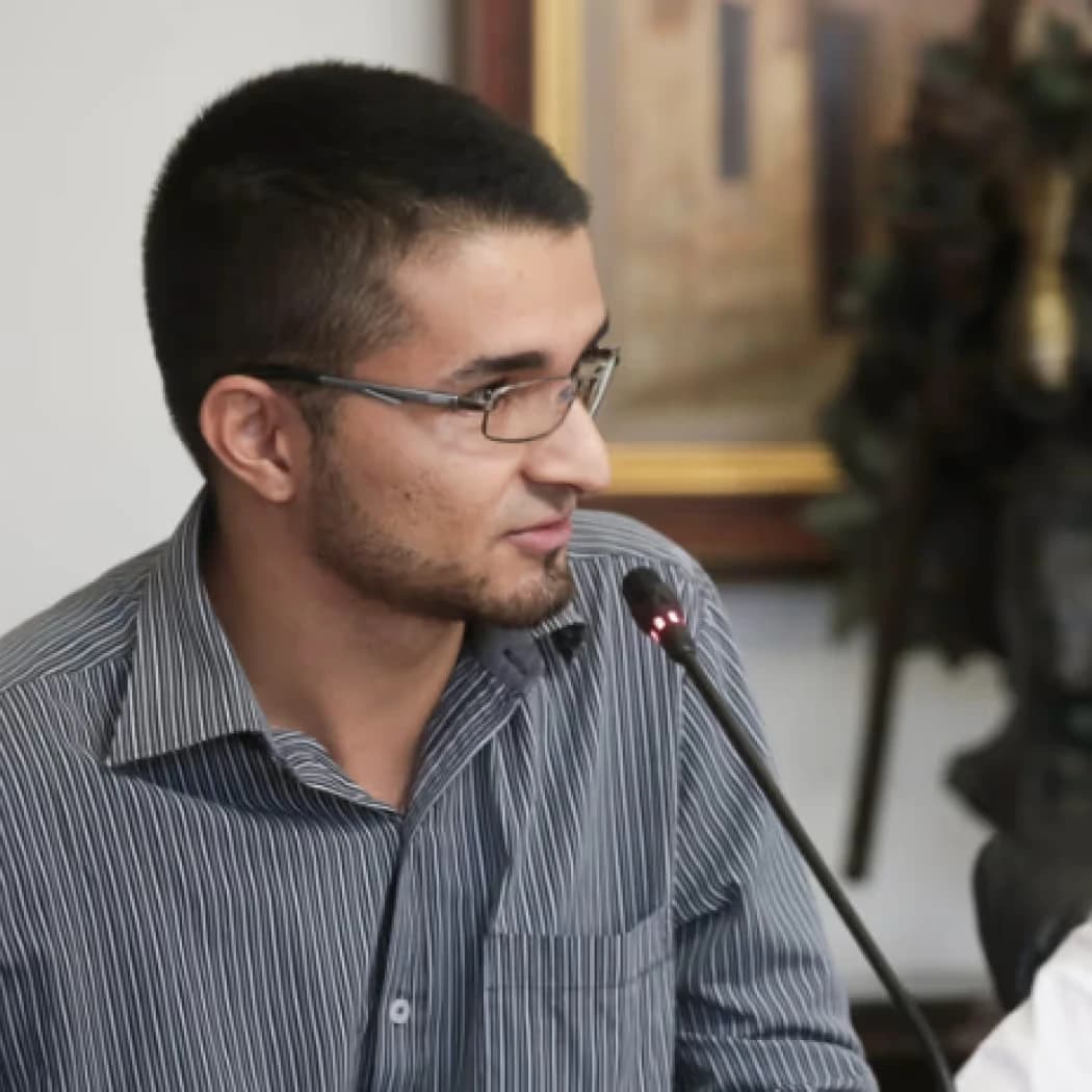 Styven speaking into a microphone at a panel discussion, wearing glasses and a striped shirt.